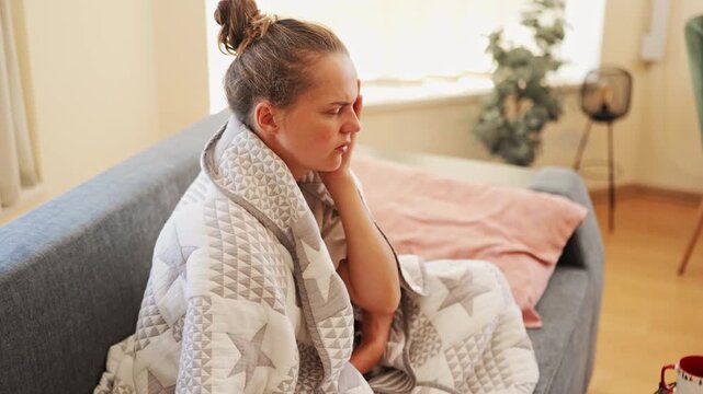 A woman is seated on a sofa with a blanket around her. She looks uncomfortable, holding her head with her eyes closed, indicating she is not feeling well