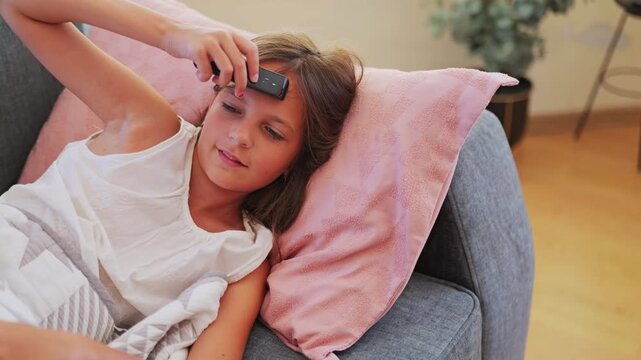 A child is lying on a couch at home, propped up by pillows. She is using a thermometer on her forehead. The room has warm lighting and light-colored decor