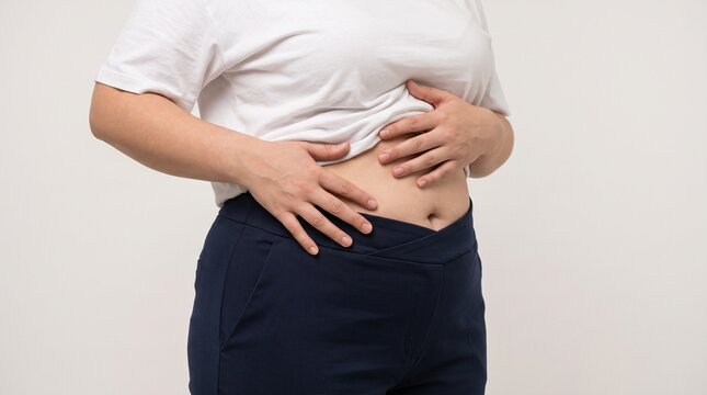 Overweight woman wearing casual white t shirt and dark blue trousers touching her chubby belly with both hands while standing indoors