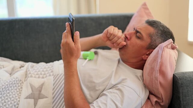A man lies on a couch with a blanket while using a smartphone. He has a hand near his mouth as if stifling a cough. The setting is indoors