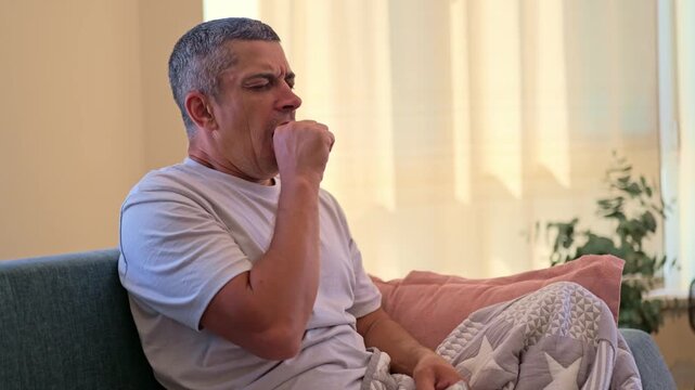A man is sitting on a couch in a bright living room. He appears to be yawning. There are plants and pillows nearby. The sunlight is coming through the window