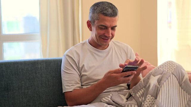 A man sits on a couch in a well-lit room. He looks happy while using his smartphone. A soft blanket covers his lap as he relaxes at home