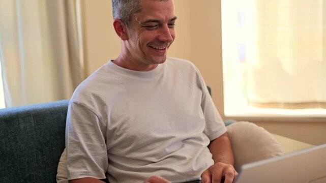 A man sits on a couch in a bright room and uses a laptop. He appears focused and engaged in his work. Soft light fills the space from nearby windows