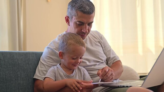 A man and a young boy are seated on a couch in a sunny room. The man interacts with a tablet while the boy watches closely. A laptop is on a nearby table