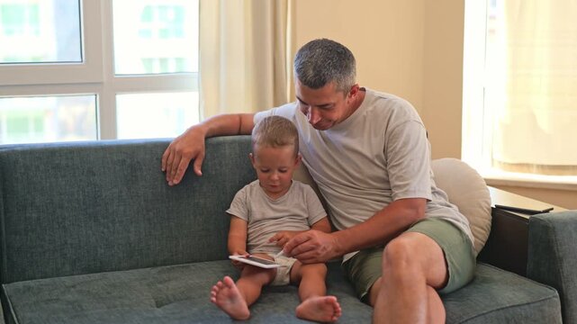 A father and his young child sit closely on a couch in a living room. They read a book together, sharing smiles and conversation as the sun shines through the windows