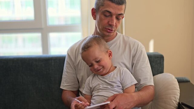 A man sits on a couch with a young boy in his lap. They are reading together, enjoying their time in a bright, quiet room at home during daylight