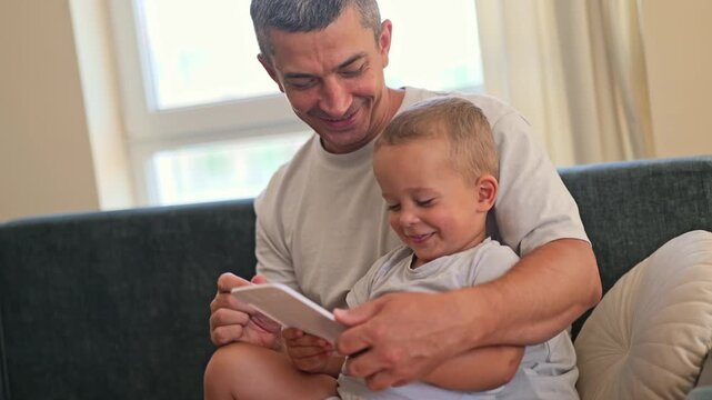 A father and his young child sit together on a sofa. They look at a smartphone, sharing a moment of joy and connection in their home