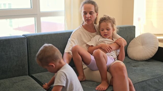 A mother sits on the couch in a living room while holding one child and watching another child play nearby. Sunlight comes through the window