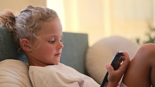 A young child is sitting on a couch with cushions, looking at a smartphone in her hands. The light coming from the window shows it is afternoon