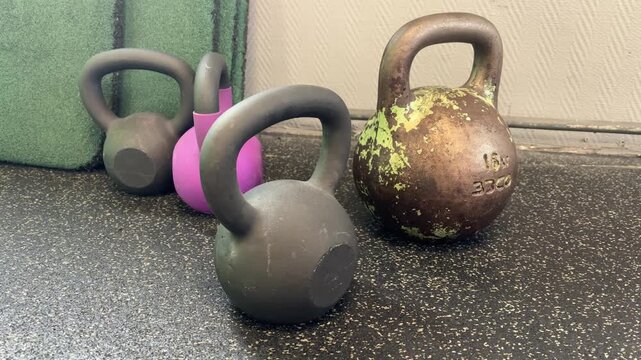 Gray and pink kettlebells stand beside a larger kettlebell with chipped green paint on a gym floor, strength equipment, functional fitness, workout preparation.