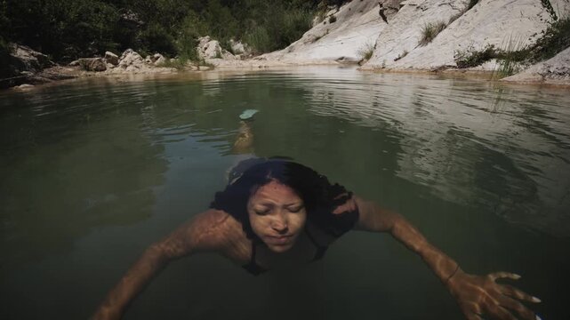 A Latina African American woman swims beneath the surface of a natural river pool, then rises out of the water near a rocky shore.