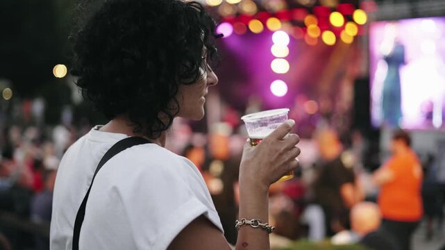 A woman is holding a cup of beer while standing at concert. Concept of enjoyment and relaxation, as the woman is likely at a social event or festival. The crowd around her suggests a lively atmosphere