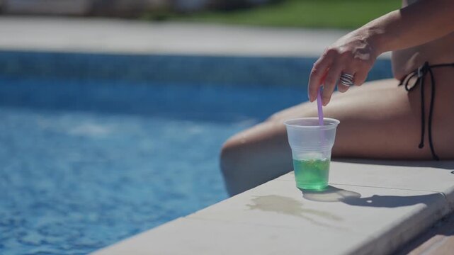 A woman is sitting on a pool deck with a green mojito cocktail or lemonade. She is holding a straw in the cup