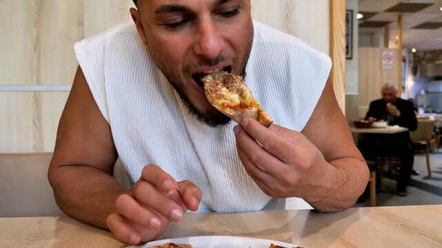 Hungry Young Athletic Man Biting into Hot Slice of Fresh Baked Tuna Pizza at Italian Restaurant Table Fast Casual Food Concept