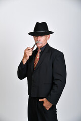 Naklejka premium Charakterporträt eines älteren Mannes im schwarzen Anzug mit Hut und Pfeife vor neutralem Hintergrund. Gentleman, Vintage-Stil, Noir-Ästhetik, Persönlichkeit und klassische Studiofotografie.