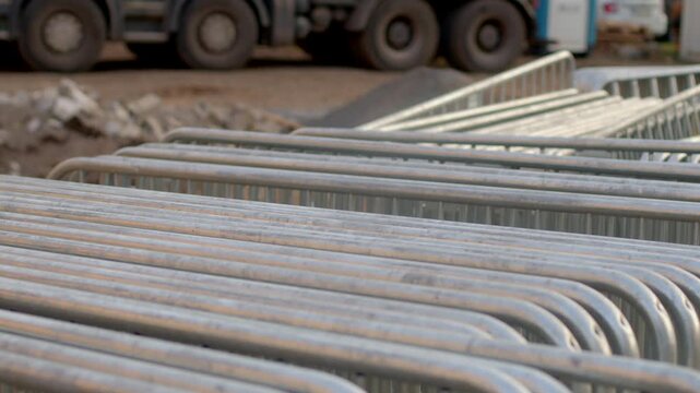 Rows of stacked metal crowd control barriers outdoors near parked vehicles, showing repeating curved tops and tubular galvanized steel surfaces for event storage and setup.