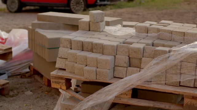 Concrete paving blocks and larger slabs are stacked on wooden pallets at outdoor construction staging area, with neat rows and daylight.
