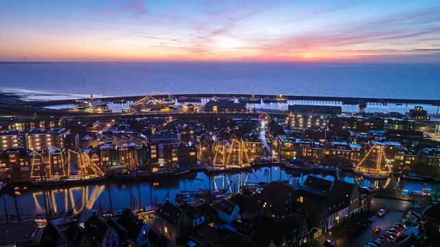 Aerial view of the historic harbor of Harlingen at twilight, featuring illuminated traditional sailing ships, charming Dutch architecture, and calm waters in Harlingen, Friesland, Netherlands.