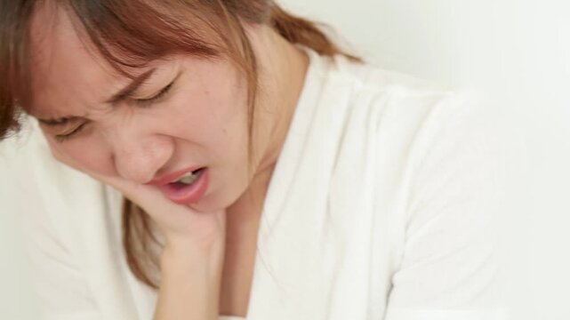 Sick 30s adult southeast asian woman having toothache, tooth decay, holding her cheek in pain with a grimace, painful facial expression, closeup shot