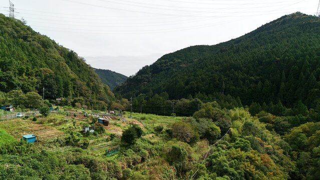 Aerial view of the upper reaches of the Akigawa River in autumn, featuring lush forested hills, small agricultural plots, and power lines in Akiruno, Tokyo, Japan.