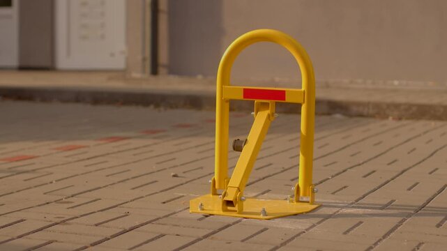 Yellow metal parking bollard stands upright on paved sidewalk, showing reflective strip and bolts at base to block reserved vehicle access.