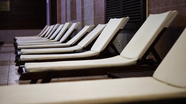 A row of white lounge chair beds are lined up in a row at hotel indoor spa. The chairs are empty and appear to be in a public area