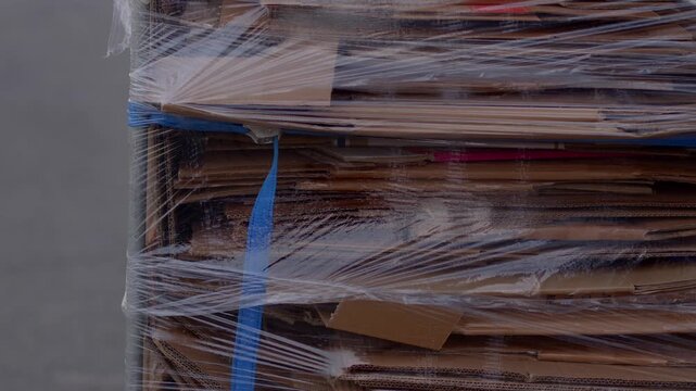 Outdoor loading area scene with pallet holding flattened cardboard boxes bundled and tightly wrapped in clear plastic stretch, secured with blue strap under overcast daylight.