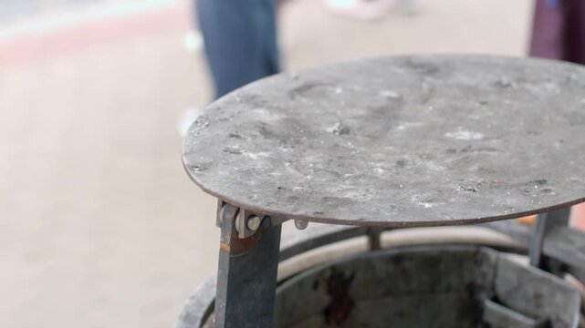 Rusty metal trash bin lid raised on hinge in outdoor school graduation celebration, with blurred students and overcast daylight in background.