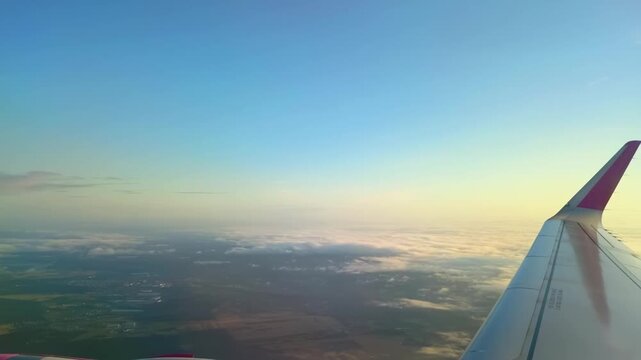 Airplane Wing Flying Above Earth and Clouds. View from airplane window of jet wing flying high above landscape terrain and white clouds under clear blue sky during flight.