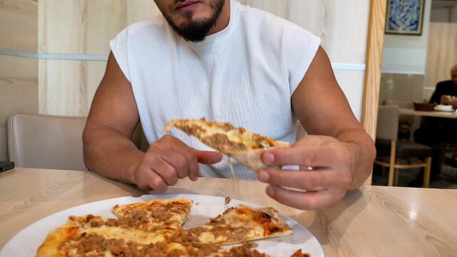 Hungry Young Athletic Man Biting into Hot Slice of Fresh Baked Tuna Pizza at Italian Restaurant Table Fast Casual Food Concept