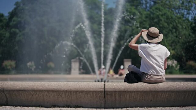 A woman wearing a straw hat sits at the fountain in Botanical garden in Cologne with water spraying in slow motion creating a cool and refreshing atmosphere. The woman is enjoying the moment