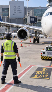 An airport ground crew worker wearing a high visibility vest holds orange marshalling wands beside a passenger aircraft, with a pushback tractor nearby.