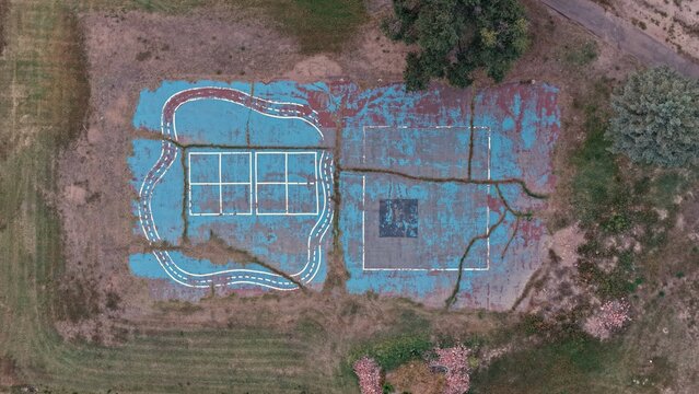 Aerial view of an old, cracked playground surface with faded blue paint, white markings, and weeds growing through the fissures in Greeley, Colorado, United States.