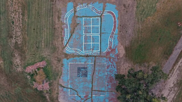 Aerial view of a weathered, cracked blue playground surface with painted white lines and a track pattern, surrounded by dry grass in Greeley, Colorado, United States.