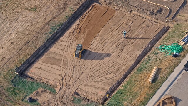 Aerial view of a skid steer loader leveling dirt at a construction site with a worker standing nearby and green pipes on the ground in Eaton, Colorado, United States.