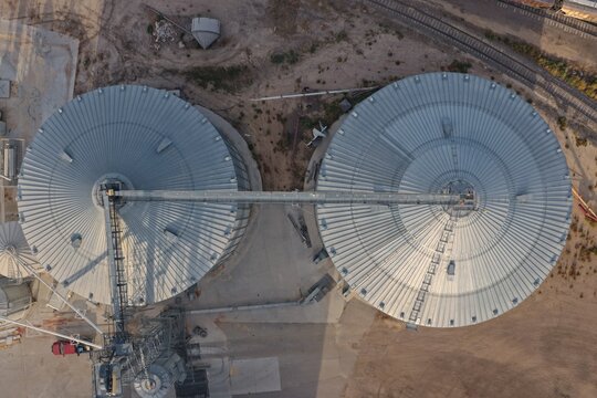 Aerial view of two large metallic grain silos connected by a catwalk, situated near a railway track in Eaton, Colorado, United States.