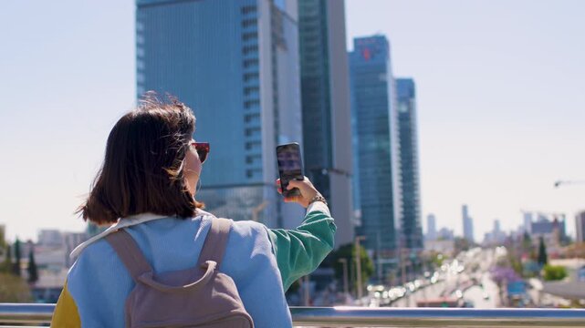 The stylish woman holds a mobile phone in her hands to photograph modern architecture in Israel. A walk on Tel Aviv. From a low angle. Travel to Israel. A modern city. 