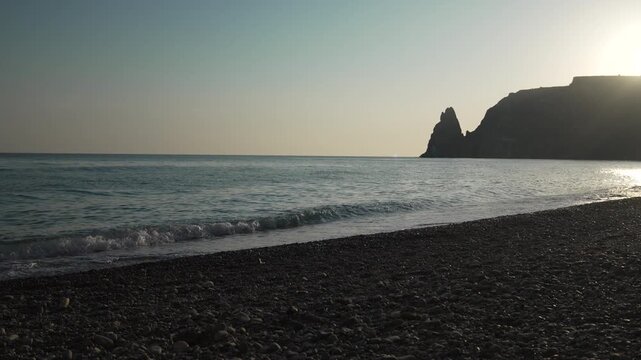 Ocean beach sunset featuring gentle waves rolling onto a dark pebble shore with a dramatic mountain silhouette against the glowing horizon