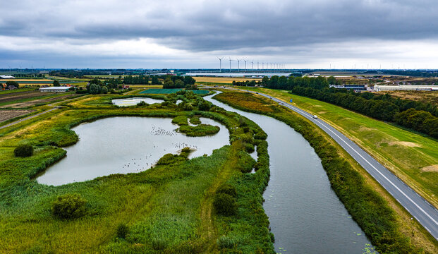 Aerial view of Agriport A7 featuring a winding canal and natural ponds surrounded by green fields and wind turbines in the distance in Middenmeer, Noord-Holland, Netherlands.