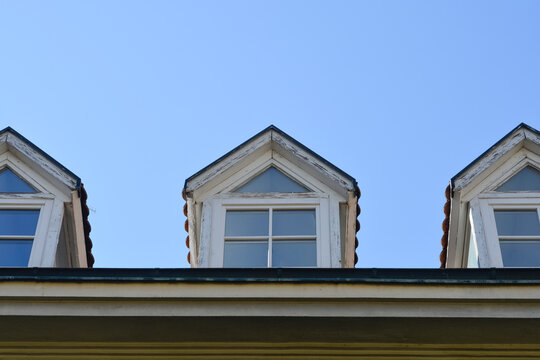 A low angle view of three old weathered wooden dormer windows with triangular tops built into a roof against a clear blue sky.