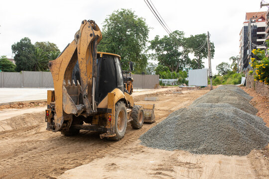 Yellow backhoe loader on road construction site with pile of crushed stone gravel, sandy ground with tire tracks, heavy machinery, earthmoving work, building under development, cloudy day
