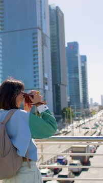 vertical shot. The girl Tourist takes a photo of the cityscape of Tel Aviv. A tourist with a camera wears a colorful jacket and backpack. Travel around Israel. modern city. 