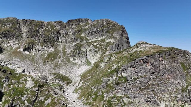 Steep rocky slopes and cliffs of Malyovitsa Peak rise above the Elenino Lake cirque under a clear blue summer sky, filmed by a moving drone.