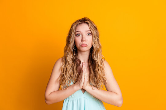 Young woman pleading with hands together in prayerful pose asking for forgiveness with hopeful expression on bright orange background