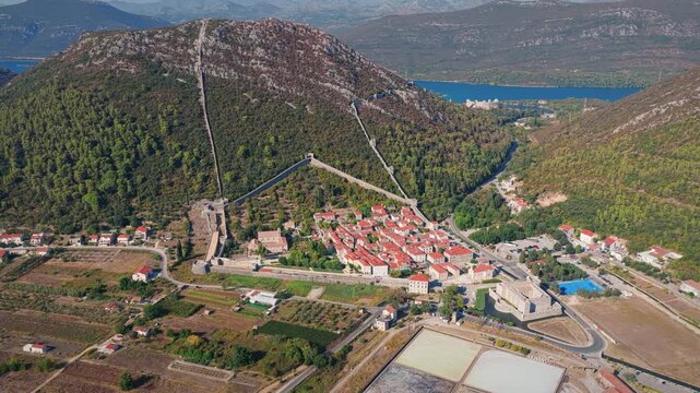 Drone shot reveals the geometric Ston Salt Pans, picturesque red-roofed village, and historic fortress walls, set between lush hills and blue waters, under clear daylight skies.