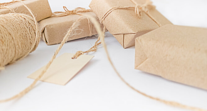 Close-up of gift boxes wrapped in brown craft paper tied with twine and blank tag on white background, concept of eco friendly holiday packaging and zero waste celebration