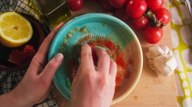 Hands grating fresh tomato on a traditional Spanish ceramic grater plate with olive oil, garlic and ripe tomatoes. Preparing authentic Mediterranean tomato spread for homemade cuisine. High quality 4k