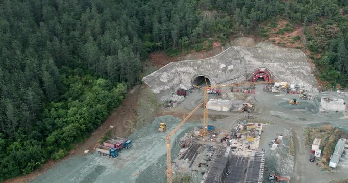 A construction site with a bridge and tunnel digging in the background. The bridge is being built over a river. The construction site is surrounded by trees and the sky is blue