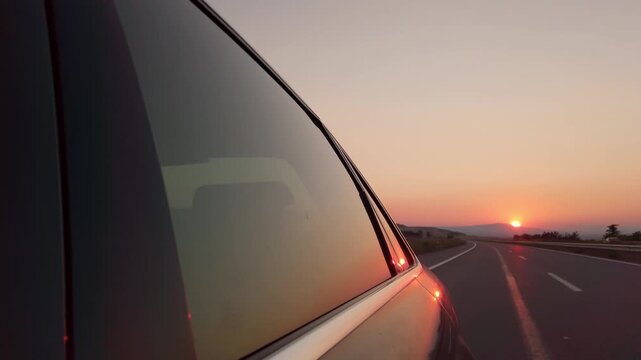 A car is driving down a road with a beautiful sunset in the background. The sun is setting, casting a warm glow over the landscape. The car is moving along the road