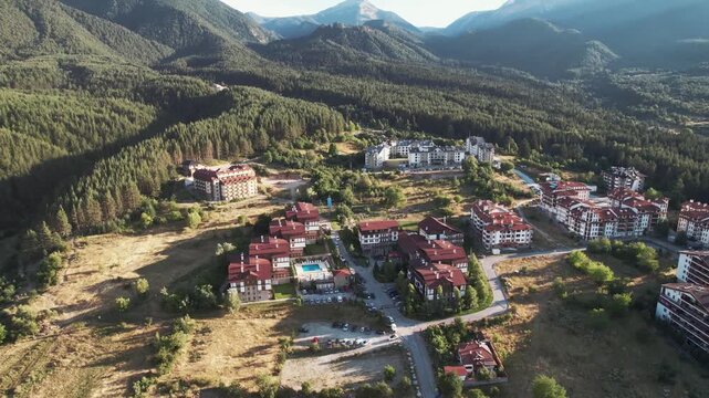 A small town with a hotel resort buildings and a pool near mountain forrest at sunset, aerial shot. The town is surrounded by mountains